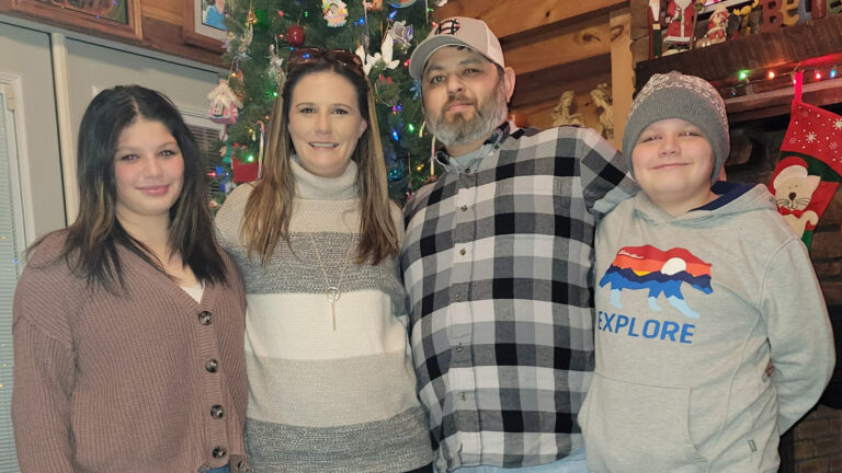 A family of four smiling together in front of a festive christmas tree and holiday decorations.