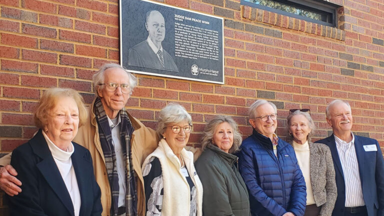 Group of smiling people gathered to honor Judge Winn legacy with a commemorative plaque on the wall behind them.