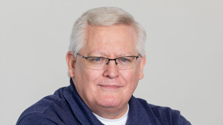 Portrait of CEO Scott Merritt, a smiling man with glasses, silver hair, and a blue shirt against a neutral background.